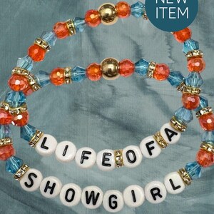 Puede incluir: Dos pulseras de cuentas con las palabras "LIFE OF A SHOWGIRL". Las pulseras presentan cuentas naranjas, azules y doradas. Un c&iacute;rculo azul con las palabras "NEW ITEM" est&aacute; en la esquina superior derecha.