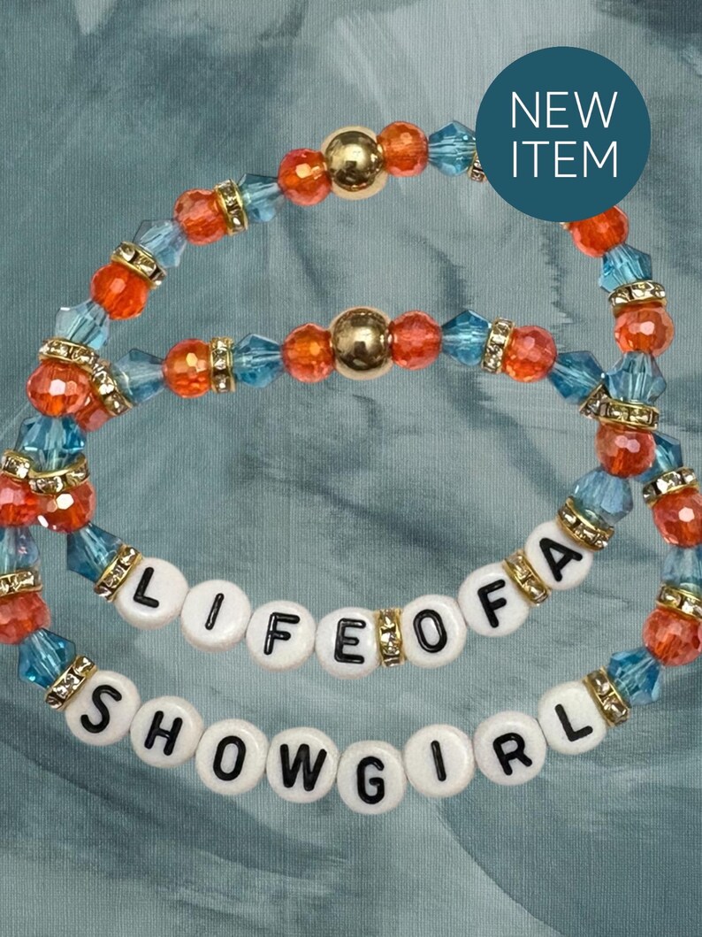 Puede incluir: Dos pulseras de cuentas con las palabras "LIFE OF A SHOWGIRL". Las pulseras presentan cuentas naranjas, azules y doradas. Un c&iacute;rculo azul con las palabras "NEW ITEM" est&aacute; en la esquina superior derecha.