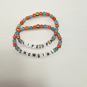 Puede incluir: Dos pulseras de cuentas. Una pulsera dice "LIFE OF A" y la otra "SHOWGIRL". Las pulseras est&aacute;n hechas de cuentas azules, naranjas y doradas. Las pulseras est&aacute;n sobre un fondo blanco.