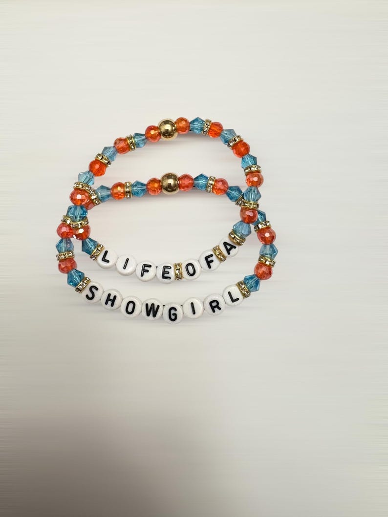 Puede incluir: Dos pulseras de cuentas. Una pulsera dice "LIFE OF A" y la otra "SHOWGIRL". Las pulseras est&aacute;n hechas de cuentas azules, naranjas y doradas. Las pulseras est&aacute;n sobre un fondo blanco.