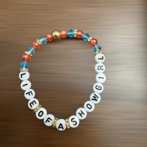 May include: A bracelet with white letter beads spelling "LIFE OF A SHOWGIRL". The bracelet has blue, orange, and gold beads, with gold accents. The background is brown.