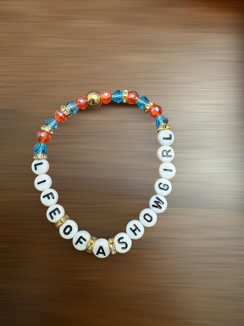 May include: A bracelet with white letter beads spelling "LIFE OF A SHOWGIRL". The bracelet has blue, orange, and gold beads, with gold accents. The background is brown.