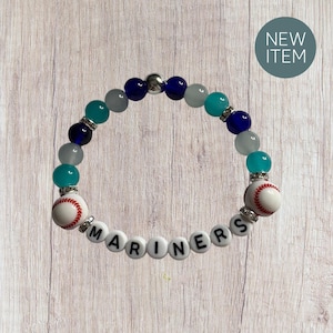 May include: A beaded bracelet featuring a baseball theme. The bracelet includes white beads with the word "MARINERS", baseball-shaped beads, and various blue, teal, and gray beads. A "NEW ITEM" sticker is in the background.