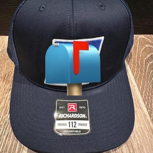 Puede incluir: Gorra de camionero azul marino con un diseño de buzón azul y una bandera roja. La gorra tiene una parte trasera de malla y una visera curva. Etiqueta de la marca Richardson. Ajustable.