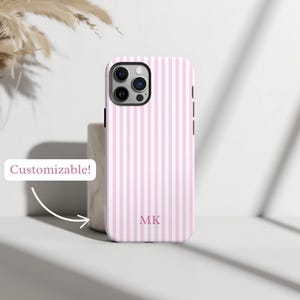 Puede incluir: Una funda de teléfono a rayas rosas y blancas con las iniciales "MK" en la parte inferior. La funda tiene un recorte para la cámara y botones laterales negros. El texto "¡Personalizable!" está en una burbuja blanca.