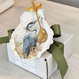 May include: An oyster shell with a heron illustration and a gold-colored spoon. The shell is mounted on a white box with a green ribbon. The text "THE CURATED FOYER" is printed on the box.