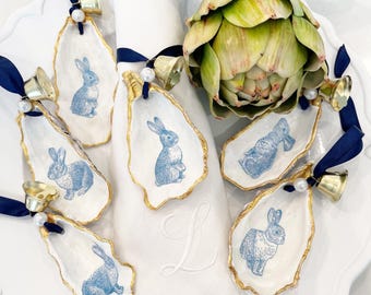 May include: A set of six oyster shell napkin rings with blue and white bunny illustrations. Each ring has a gold rim and is tied with a navy blue ribbon and a small bell.