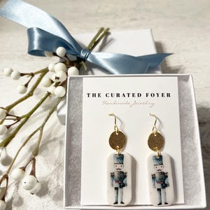 May include: A pair of gold and white porcelain earrings featuring a nutcracker design. The earrings are packaged in a white box with the text "THE CURATED FOYER Handmade Jewelry".