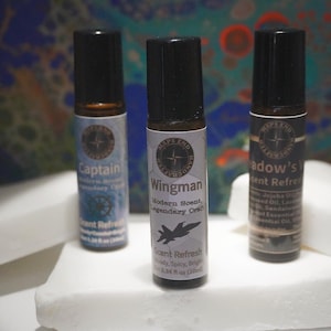May include: Three roll-on bottles of scent refreshers. The bottles have black caps and white labels with text and graphics. The labels read "Captain", "Wingman", and "Shadow's". The "Wingman" label features a jet graphic. The bottles are on a white surface.