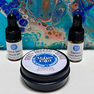 May include: An image of beard grooming products. Two small glass bottles with black droppers are labelled "Tempest Tamer Beard Oil" and "Captain Scent Refresh". A black tin of "Tradewind" pomade is in the centre. The background is an abstract painting.