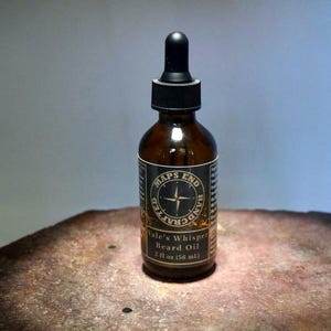 Op de afbeelding: Een bruine glazen fles baardolie met een zwarte druppelaar. Het etiket luidt "Maps End Handcrafted" en "Vale's Whisper Beard Oil, 56 ml". De fles staat op een getextureerd oppervlak.