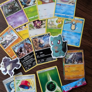 May include: A collection of Pokémon trading cards and stickers, featuring various characters like Skwovet, Wimpod, and Phantump. The cards display colorful illustrations and game information. Also included are a green energy card, a Pot Helmet trainer card, and a candy.