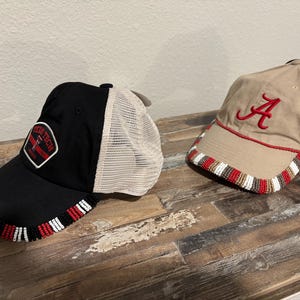 Peut inclure: Deux casquettes de baseball avec bordure perlée. L'une est noire avec un dos en filet blanc et un écusson "TECH TECH". L'autre est beige avec un "A" rouge et une bordure rouge.