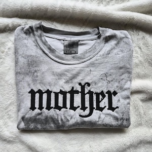 May include: A folded gray t-shirt with the word "mother" in black gothic lettering. The shirt has a marbled effect and a crew neck. The tag reads "Comfort Colors" and the size is L-G-G.