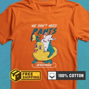 May include: Orange t-shirt with a graphic of a shark riding a yellow duck floatie. Text above reads "WE DON'T NEED PANTS" in blue and white. The word "NEVERMORE" is below the graphic. The shirt is made of 100% cotton.