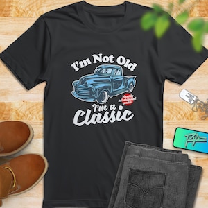 Pode incluir: T-shirt preto com um gráfico de um caminhão clássico azul e o texto "I'm Not Old, I'm a Classic." O gráfico inclui as palavras "Nearly all original parts." A camisa é exibida com jeans e sapatos marrons.