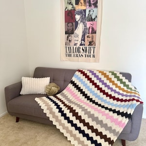 May include: A gray couch with a white pillow and a crocheted blanket with colorful stripes. The blanket has a chevron pattern. A poster on the wall behind the couch reads "Taylor Swift The Eras Tour".