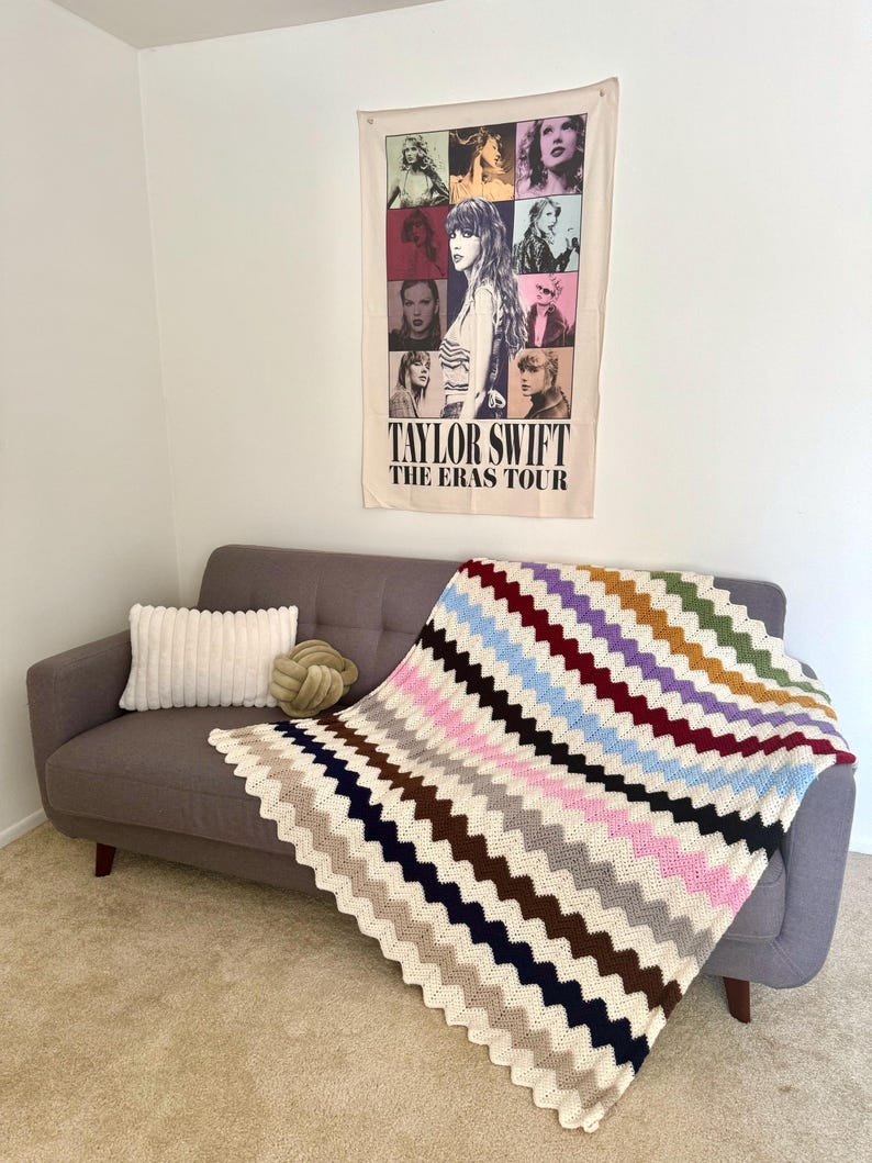 May include: A gray couch with a white pillow and a crocheted blanket with colorful stripes. The blanket has a chevron pattern. A poster on the wall behind the couch reads "Taylor Swift The Eras Tour".