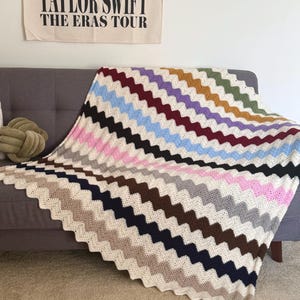 May include: A crocheted blanket with a chevron pattern in various colors, including pink, blue, brown, and gray. The blanket is draped over a gray couch.