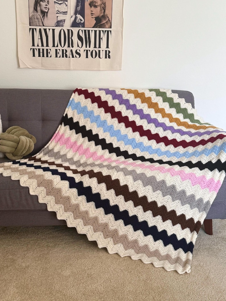 May include: A crocheted blanket with a chevron pattern in various colors, including pink, blue, brown, and gray. The blanket is draped over a gray couch.