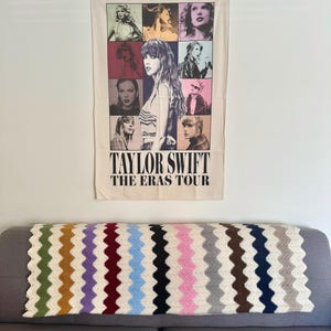 May include: A white tapestry with a collage of images of Taylor Swift in different eras. The text "Taylor Swift The Eras Tour" is printed at the bottom of the tapestry.