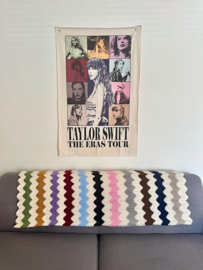 May include: A white tapestry with a collage of images of Taylor Swift in different eras. The text "Taylor Swift The Eras Tour" is printed at the bottom of the tapestry.