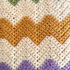 May include: A chevron patterned crocheted blanket with stripes of green, cream, mustard yellow, and purple.