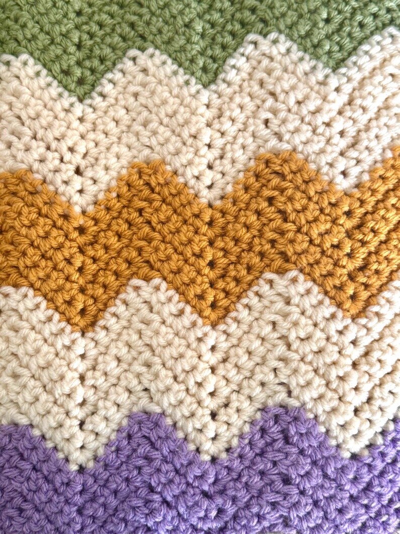 May include: A chevron patterned crocheted blanket with stripes of green, cream, mustard yellow, and purple.