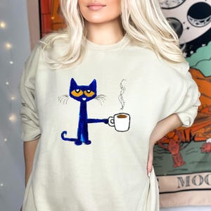 May include: A light blue sweatshirt with a blue cat holding a cup of coffee. The cat has orange eyes and a white outline. The text 'MOON' is visible in the background.