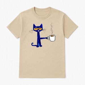 May include: A tan t-shirt with a blue cat holding a white coffee cup with a brown liquid inside. The cat has orange eyes and a black nose. The cat is drawn in a simple, childlike style.