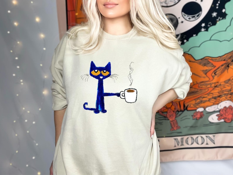 May include: A light blue sweatshirt with a blue cat holding a cup of coffee. The cat has orange eyes and a white outline. The text 'MOON' is visible in the background.
