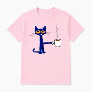 May include: Pink t-shirt with a blue cat holding a cup of coffee. The cat has orange eyes and a white cup with brown coffee.