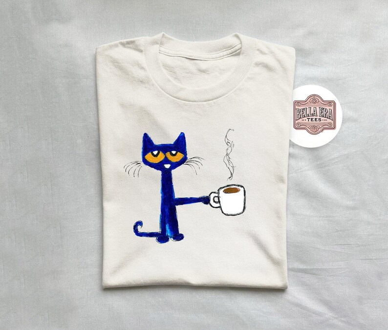 May include: White t-shirt with a blue cat holding a cup of coffee. The cat has orange eyes and a white mustache. The text "Bella Era Tees" is in a brown rectangle on the upper right corner of the shirt.