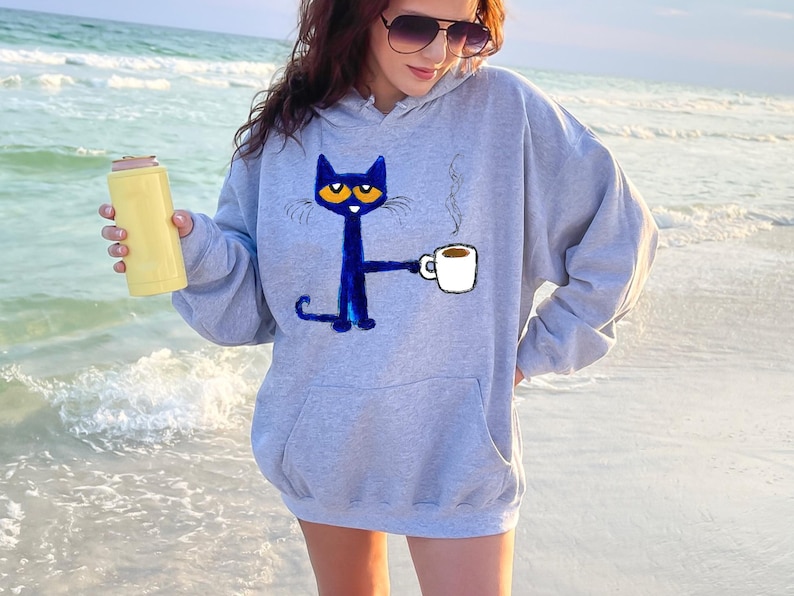May include: A gray hoodie with a blue cat holding a cup of coffee. The cat has orange eyes and a white mustache. The text "Coffee" is written on the cup.