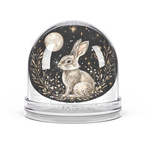 Könnte beinhalten: Eine klare Schneekugel mit einer Aquarellillustration eines Kaninchens, eines Vollmonds und Sternen vor einem schwarzen Hintergrund. Die Basis der Schneekugel ist transparent.