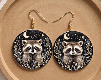 May include: A pair of round earrings featuring a raccoon design. The earrings have a black background with a crescent moon, stars, and floral accents. The raccoon is depicted in shades of brown and white, with gold-colored ear wires.