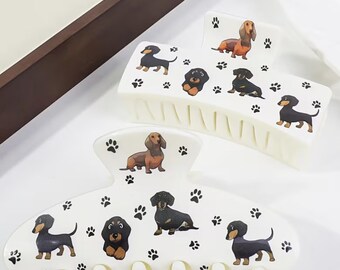 Könnte beinhalten: Zwei cremefarbene Haarklammern mit Dackel-Illustrationen und Pfotenabdrücken. Eine Klammer zeigt einen braunen und schwarzen Dackel, während die andere verschiedene Dackel-Designs zeigt. Diese Klammern sind ein lustiges Accessoire für Hundeliebhaber.