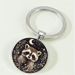 May include: A silver-toned metal keychain featuring a circular pendant with a black background. The pendant displays a brown and white raccoon illustration surrounded by gold-colored botanical line art, small stars, and a crescent moon.