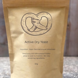 以下が含まれることがあります： 活性乾燥酵母の茶色の紙袋。プレッツェルのイラストと「Active Dry Yeast」の文字が特徴です。材料と指示も記載されています。袋には60gの酵母が入っています。