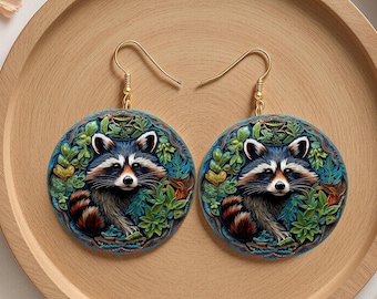 May include: A pair of round earrings featuring a raccoon design. The raccoon has a black and white face, surrounded by green foliage. The earrings have gold-colored hooks and are displayed on a wooden tray.