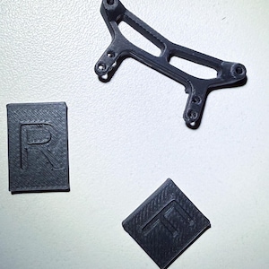 Può includere: Tre pezzi stampati in 3D di colore nero, uno è un piccolo pezzo rettangolare con la lettera "R" stampata su di esso, un altro è un piccolo pezzo a forma di diamante con la lettera "T" stampata su di esso, e il terzo è un pezzo più grande con una forma curva e diversi fori.