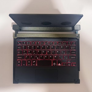 May include: A compact black wireless keyboard with a built-in trackpad and red backlit keys. The keyboard features a standard QWERTY layout with function keys and is housed within a dark, textured frame that includes a hinged, elevated stand.