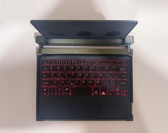 May include: A compact black wireless keyboard with a built-in trackpad and red backlit keys. The keyboard features a standard QWERTY layout with function keys and is housed within a dark, textured frame that includes a hinged, elevated stand.