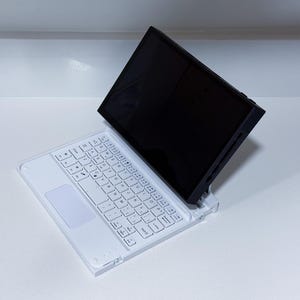 Puede incluir: Una funda de teclado blanca para tableta con un panel táctil integrado y una pantalla negra. El teclado tiene teclas blancas y está inclinado hacia arriba, lo que sugiere que está abierto y listo para usar. La configuración está sobre una superficie blanca.