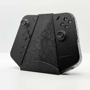 Puede incluir: Un mando de juegos negro con un soporte a juego. El mando tiene botones, un panel direccional y joysticks analógicos. El soporte tiene un patrón geométrico. Diseñado para juegos portátiles.