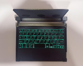 May include: A compact black wireless keyboard with a QWERTY layout and vibrant green backlit keys. The device features a built-in trackpad below the keys and a dark, textured stand attached to the top edge, designed to hold a tablet or smartphone.