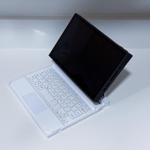 May include: A white tablet keyboard case with a built-in touchpad and a black screen. The keyboard has white keys and is angled upwards, suggesting it is in use. The setup is on a white surface.
