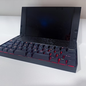 Pode incluir: Um dispositivo de computador portátil preto com um ecrã escuro e um teclado mecânico com teclas retroiluminadas a vermelho. O chassis tem um painel geométrico texturado acima das teclas e um design compacto e angular.