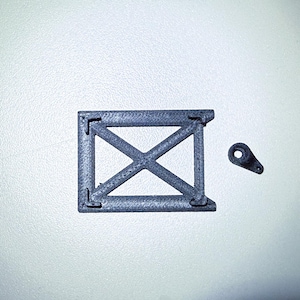 Può includere: Una cornice in plastica grigia con un design a forma di croce e un piccolo pezzo di plastica grigia con un foro al centro.