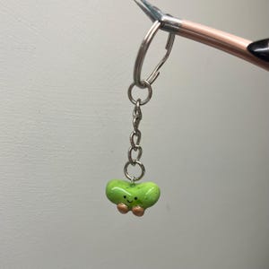 Op de afbeelding: Een groene, boonvormige sleutelhanger met een glimlachend gezicht en kleine bruine armen. De sleutelhanger is bevestigd aan een zilveren ketting en een zilveren ring.
